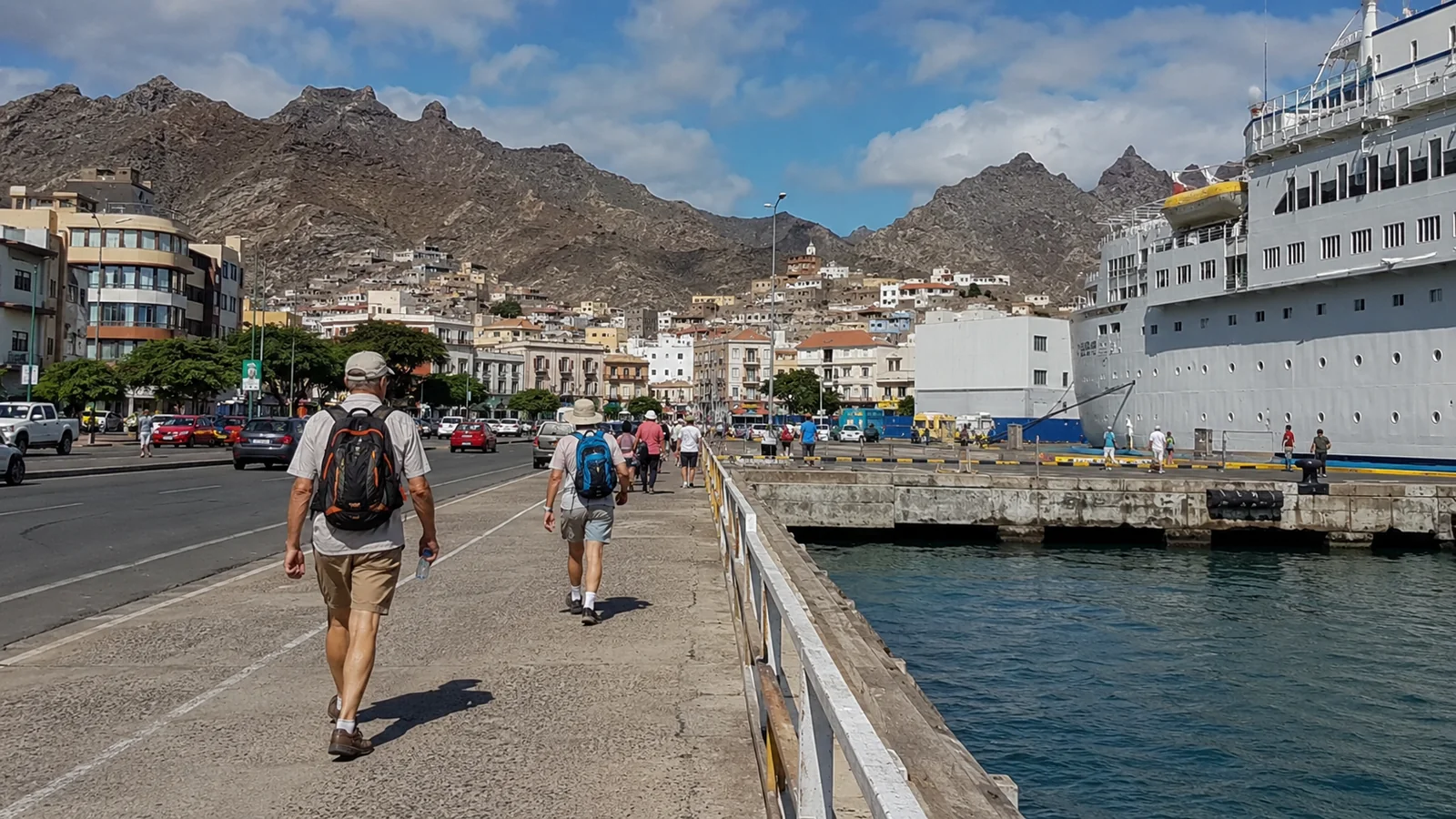 Passengers walking beside a docked cruise ship with steep mountains behind