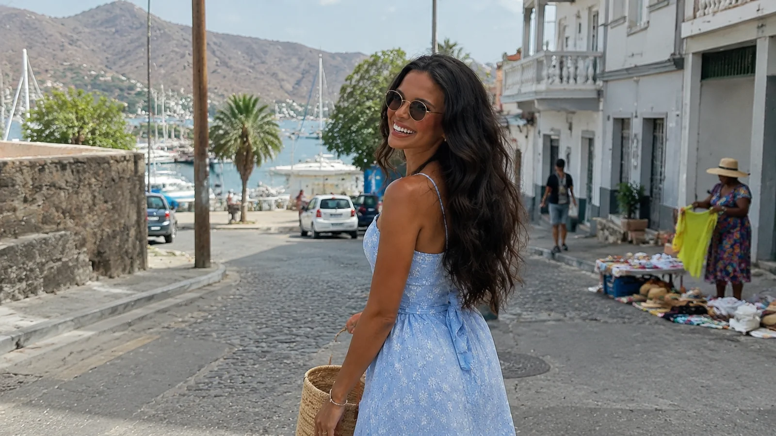 Smiling woman in a blue dress on a cobblestone street overlooking a harbor