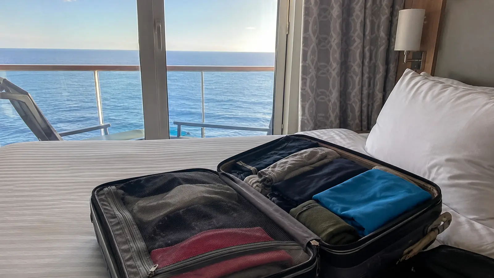 Open packed suitcase on a cruise cabin bed beside an ocean-view balcony