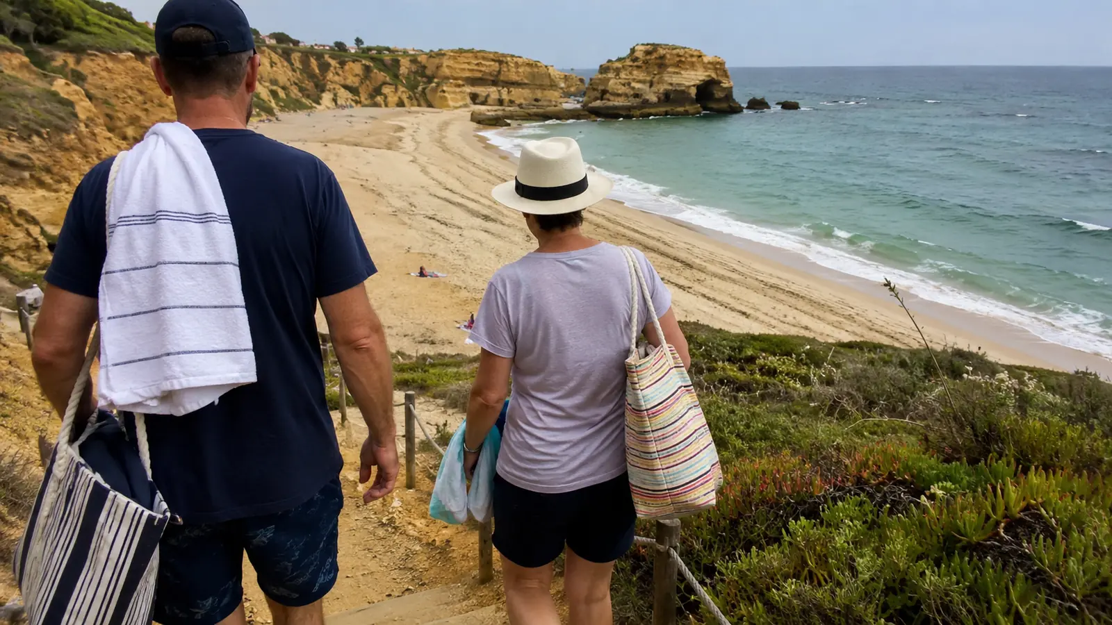 Man and woman carrying beach bags while walking along a sandy coastal path