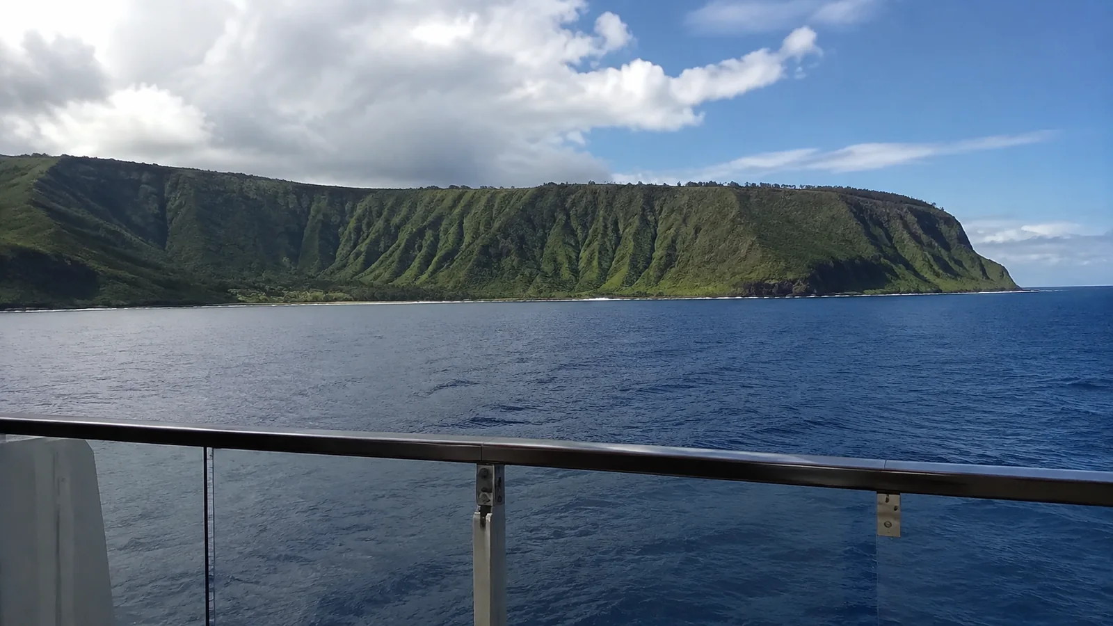 Green coastal cliffs rise beyond the ocean and a ship railing.