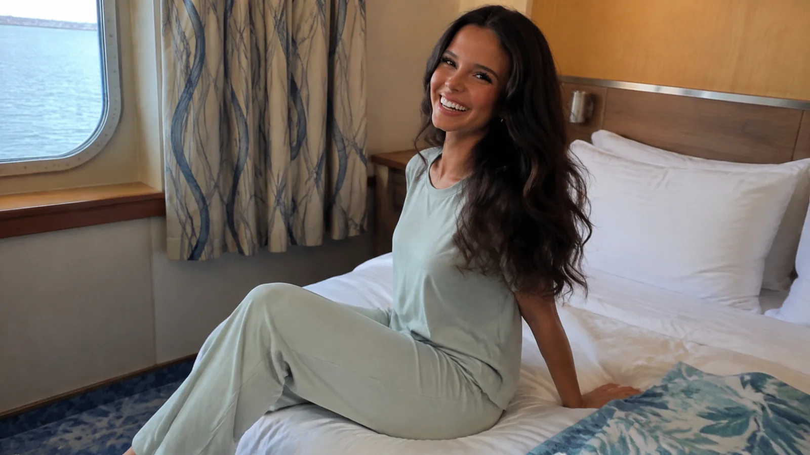 Woman sits smiling on a bed beside a stateroom window.