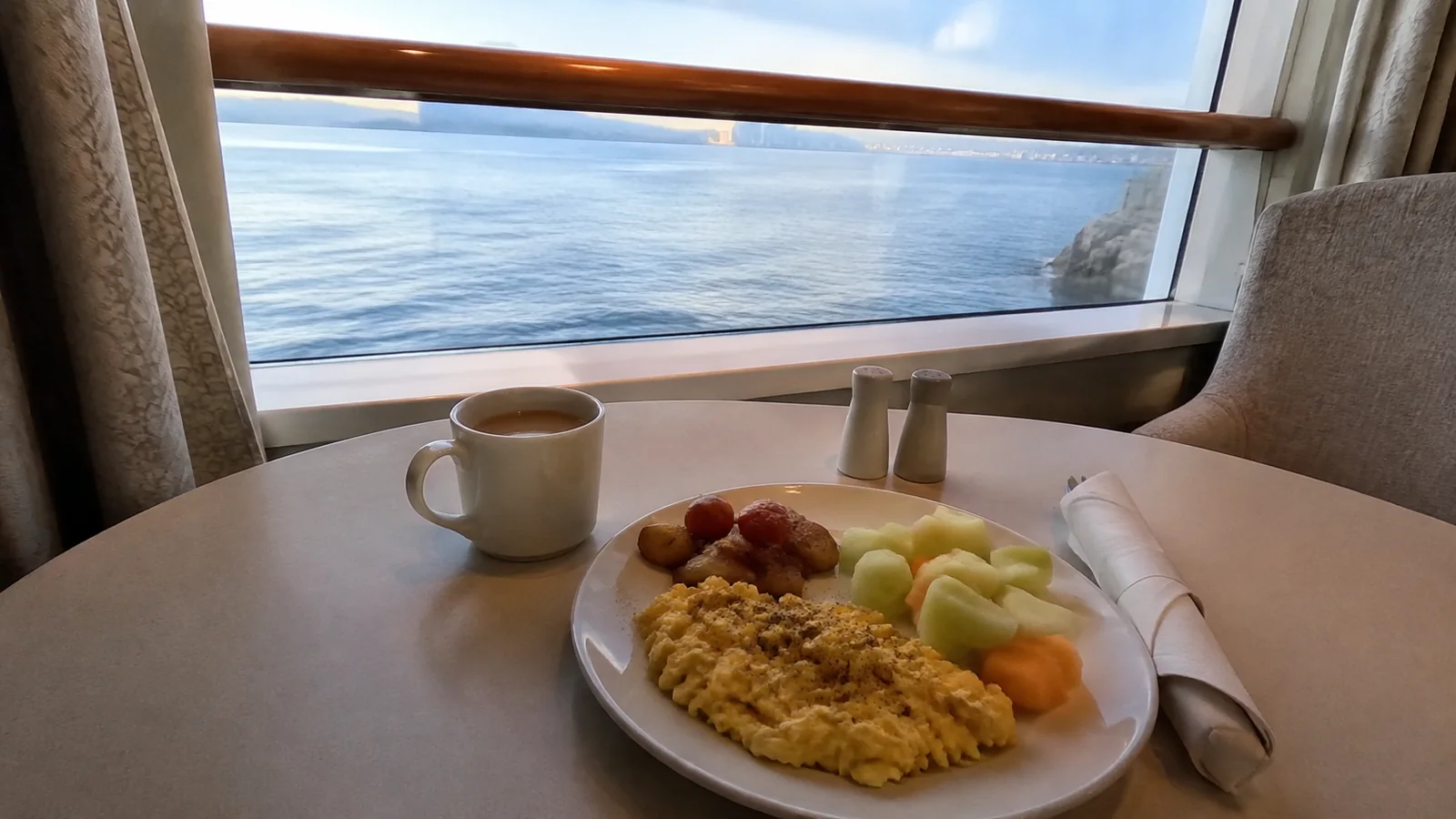 Eggs, fruit and coffee sit on a table beside an ocean window.