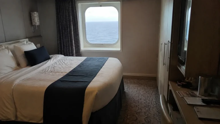 Ocean-view cruise cabin with a double bed, desk and wardrobe.
