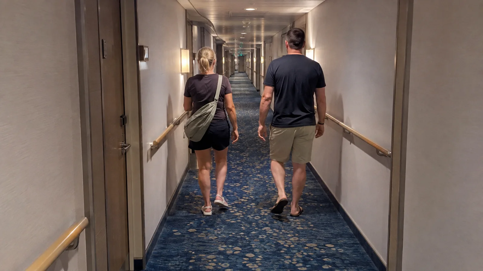 Two passengers walk along a carpeted cruise ship corridor.