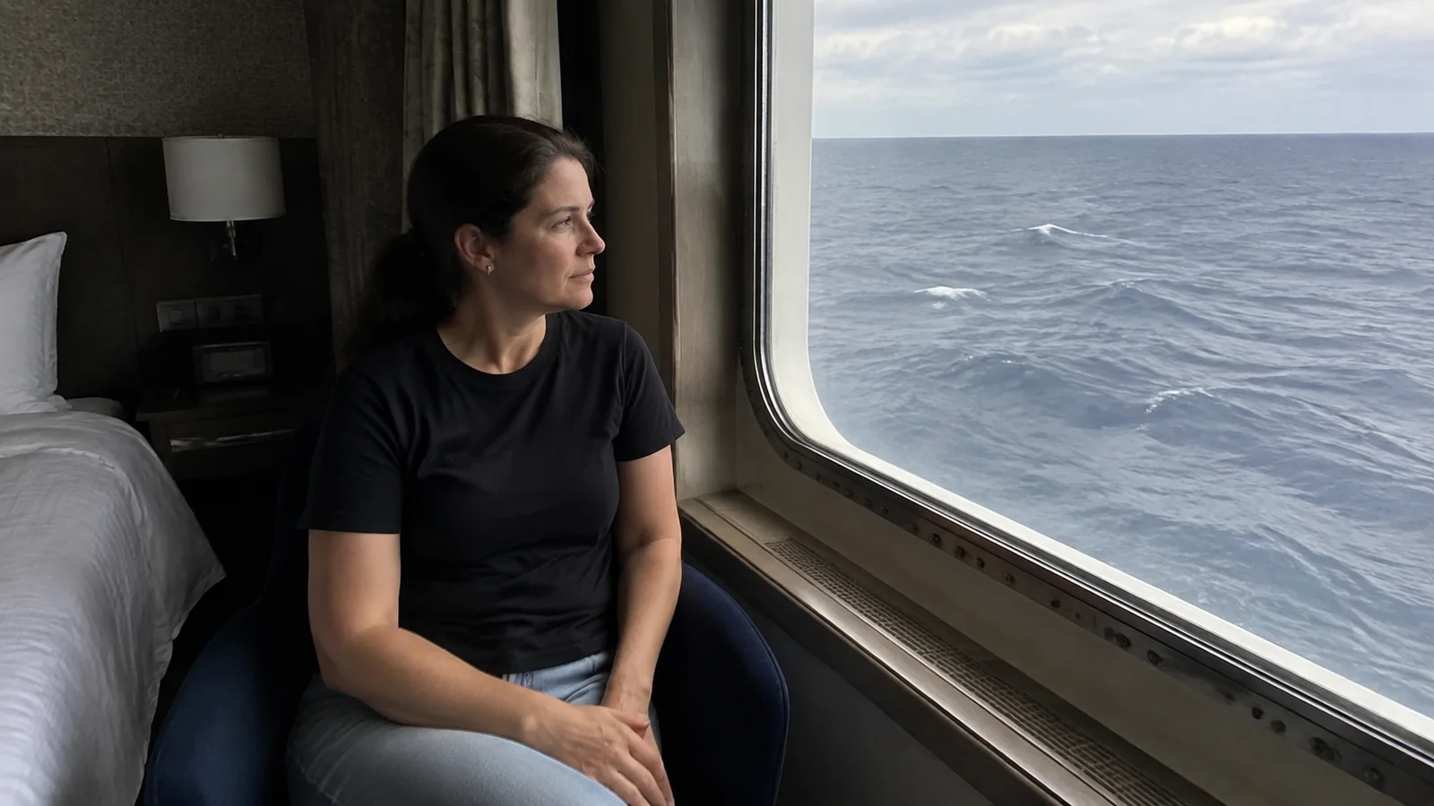 Woman sits beside a stateroom window looking at the sea.
