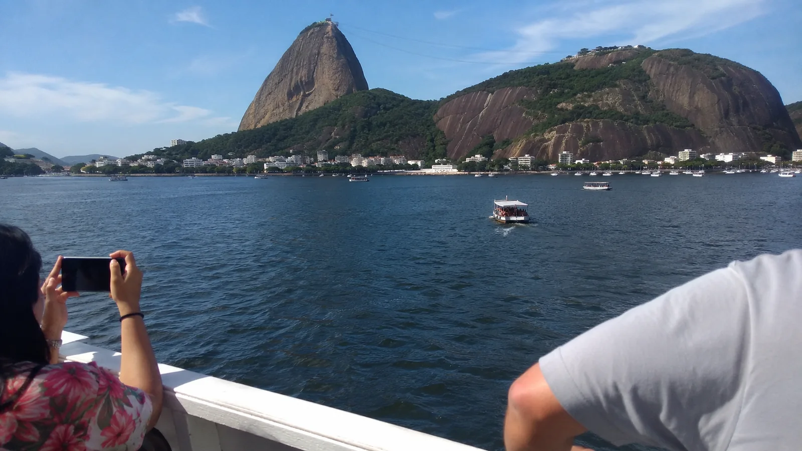 Passengers photographing a steep rounded mountain across a blue bay from a boat deck