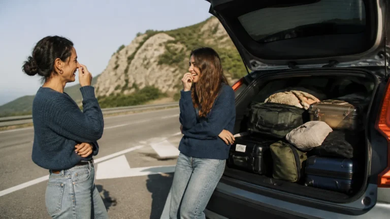 Two women laugh beside an open car trunk filled with luggage.