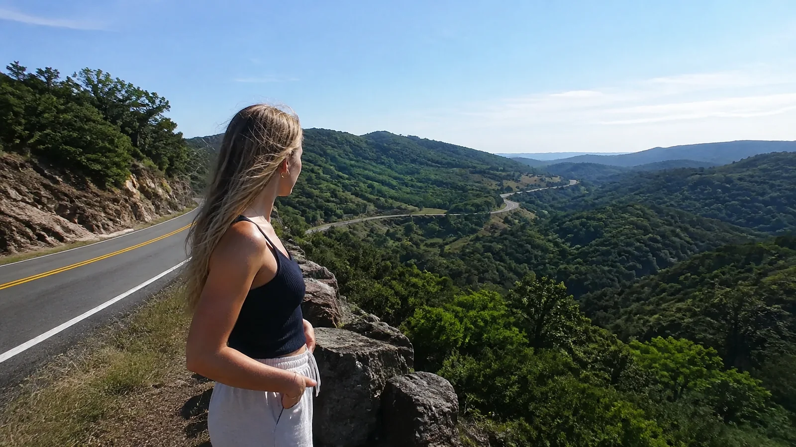 Woman stands at a roadside overlook above forested hills.