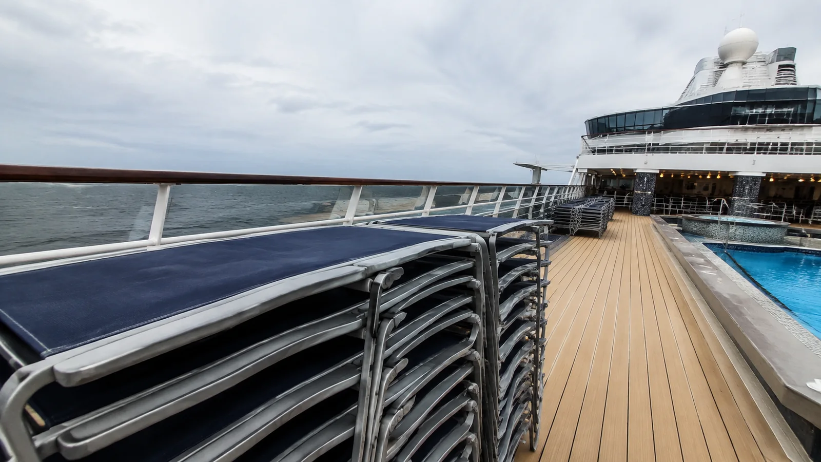 Stacked lounge chairs line a ship deck beside a swimming pool.
