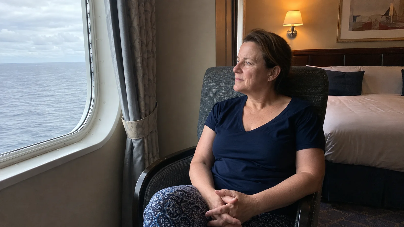 Woman sits in a stateroom chair looking through an ocean window.