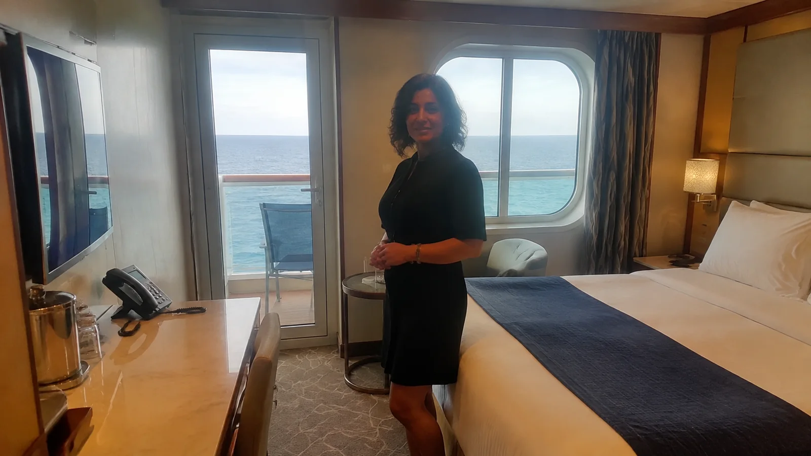 Woman stands beside a bed in a balcony stateroom.