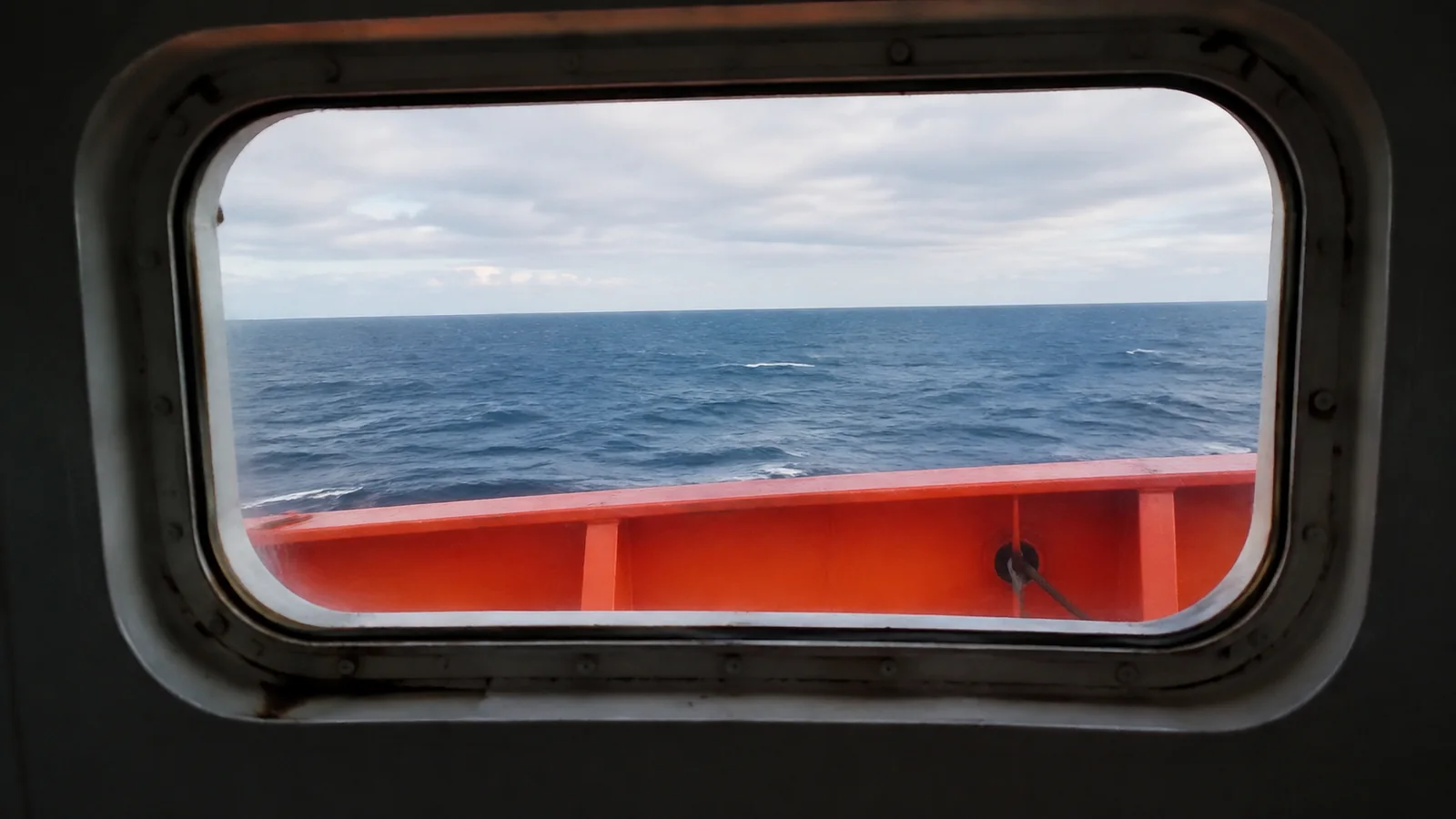 Orange lifeboat roof partially obstructs the view through a ship window.