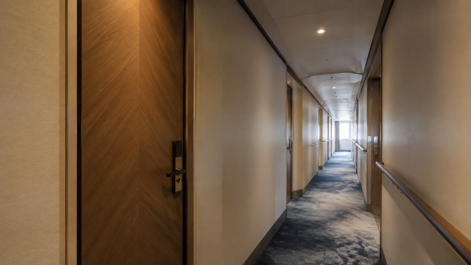 Empty carpeted stateroom corridor extends toward a bright window.