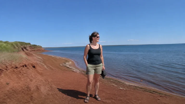 Woman standing on a red sand shoreline beside calm blue water.