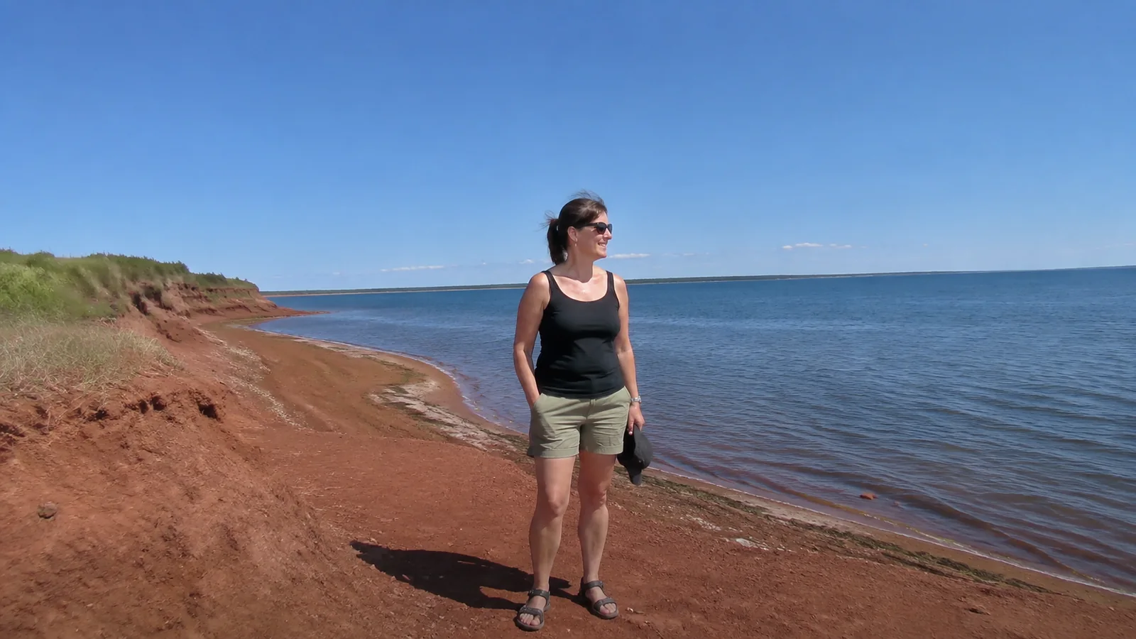 Woman standing on a red sand shoreline beside calm blue water.