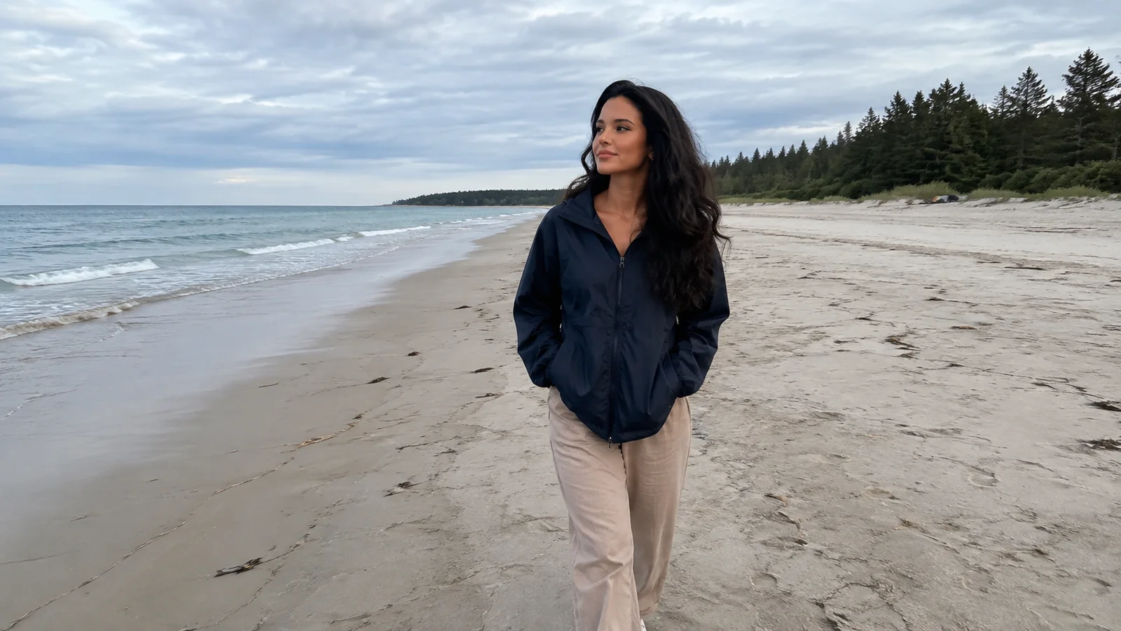 Woman walking a pale sandy beach bordered by evergreen trees.