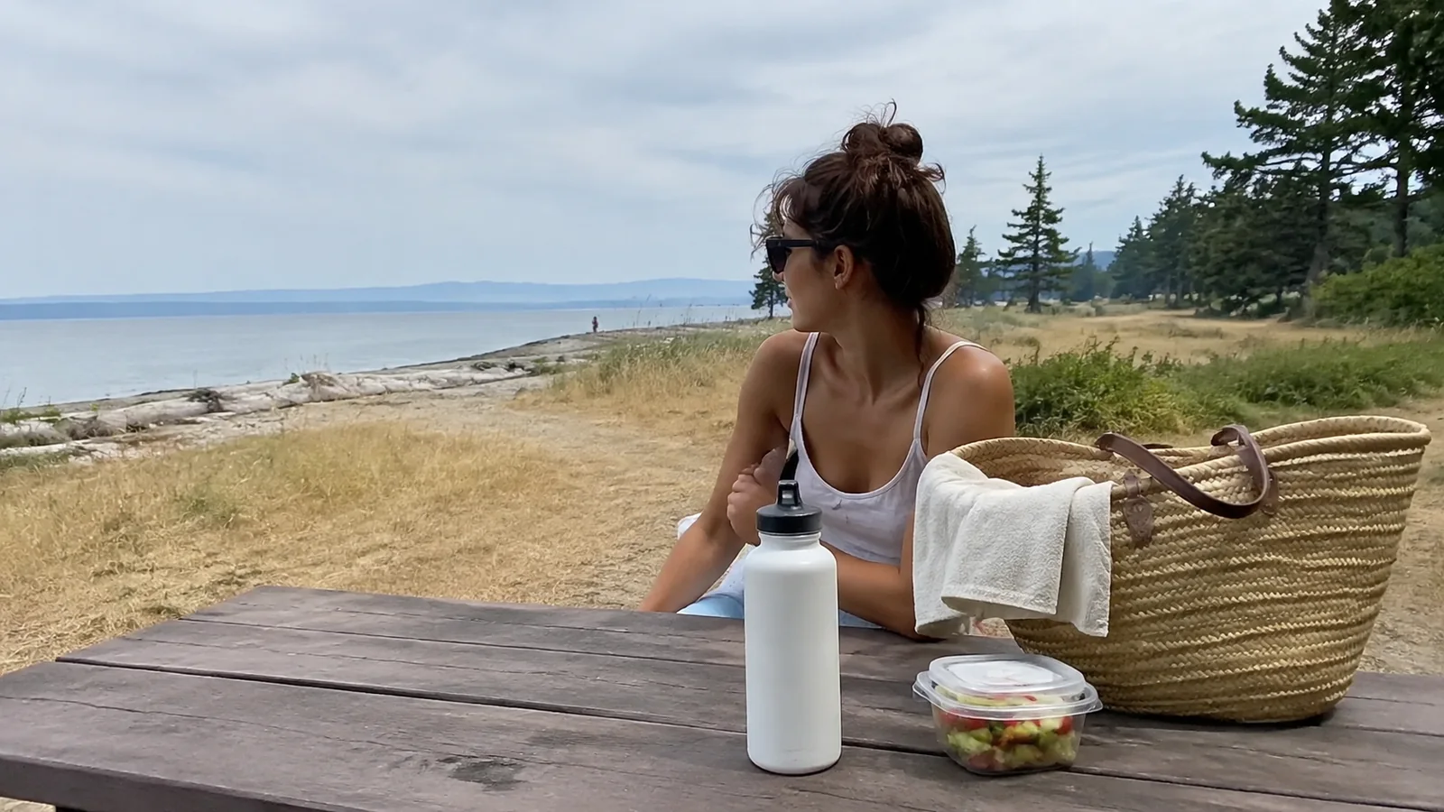 Woman seated at a coastal picnic table with a tote and water bottle.