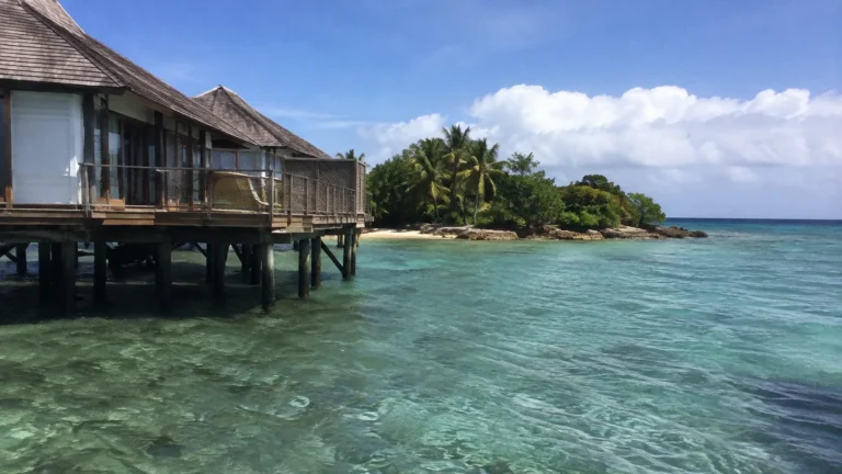 Overwater bungalow with a wooden deck beside clear shallow water.
