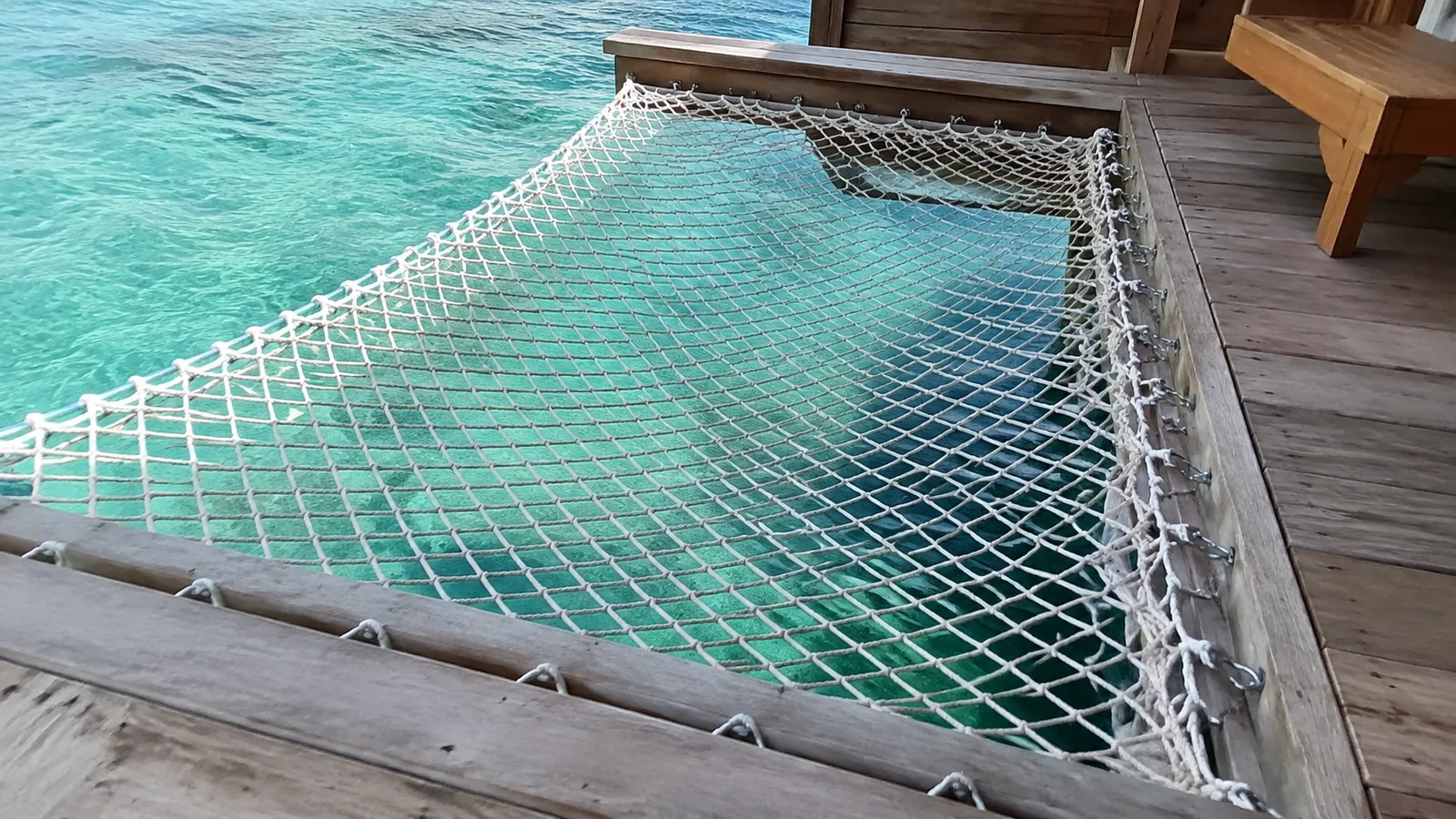 Rope hammock suspended over turquoise water beside a villa deck.