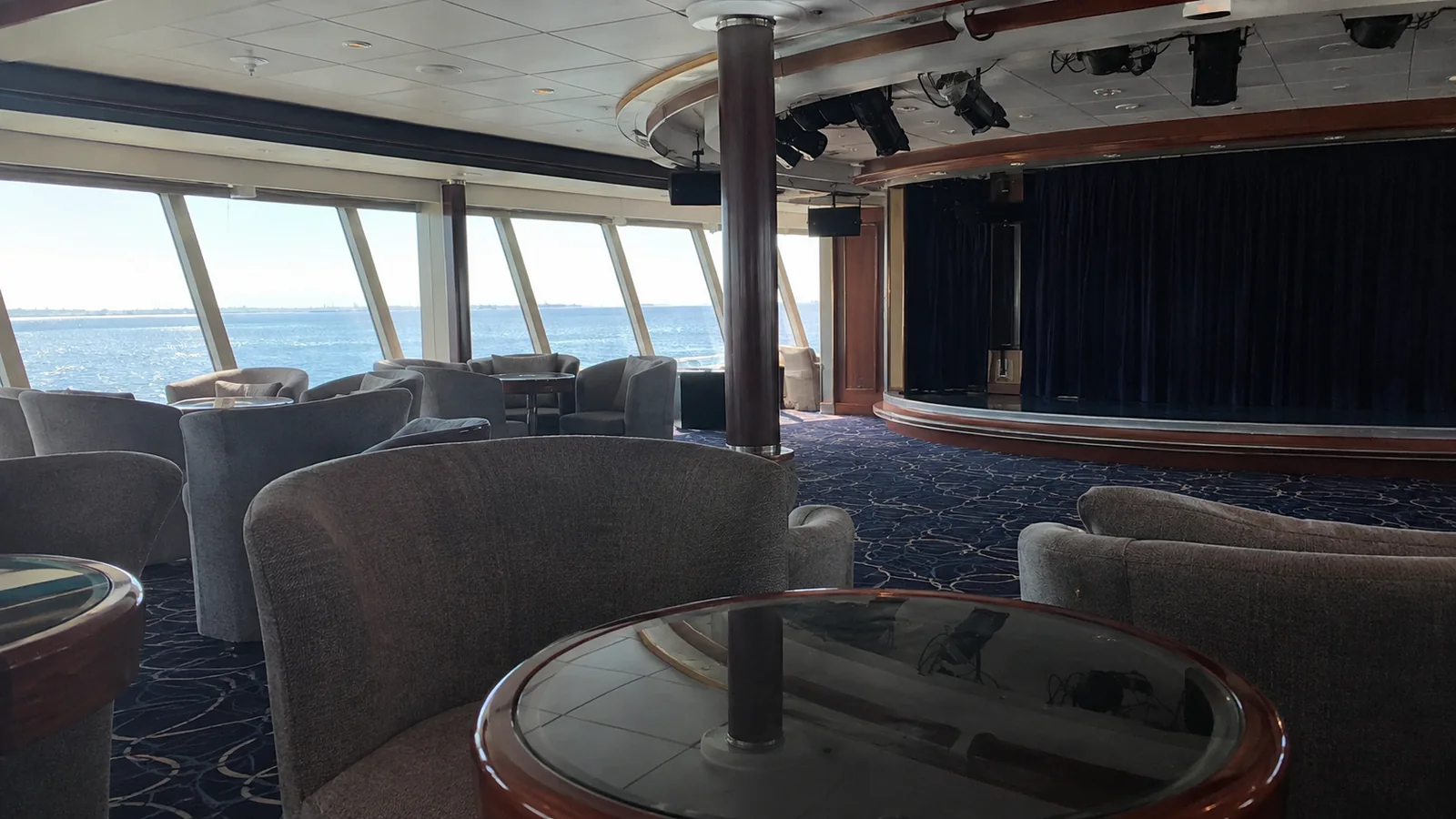 Empty lounge chairs facing a small stage beside ocean-view windows.