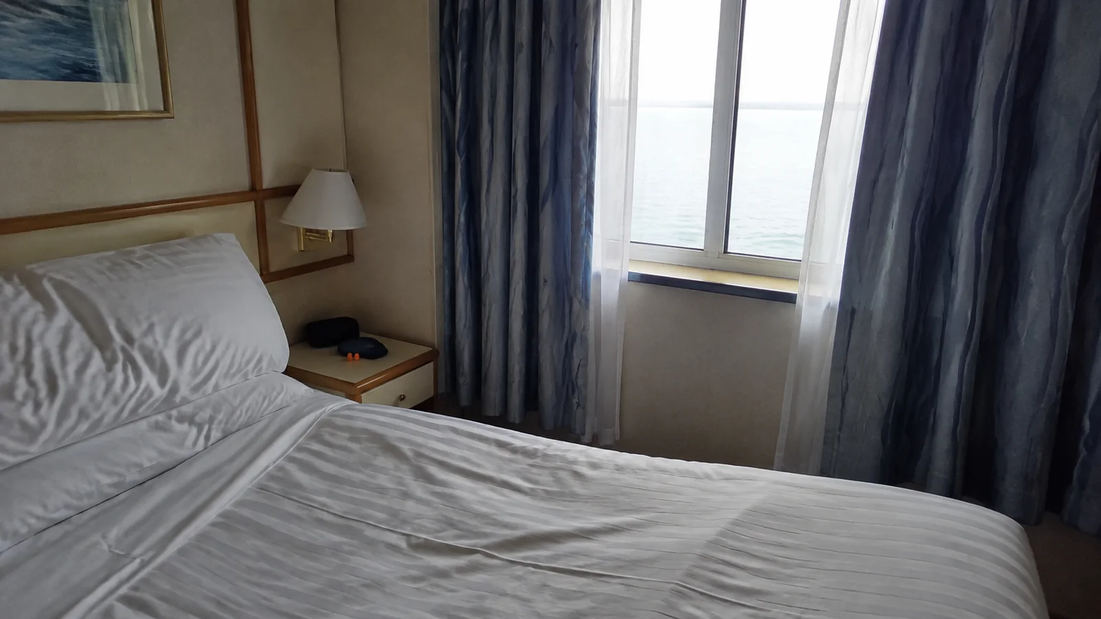 Cruise cabin bed beside thick curtains and an ocean-view window.