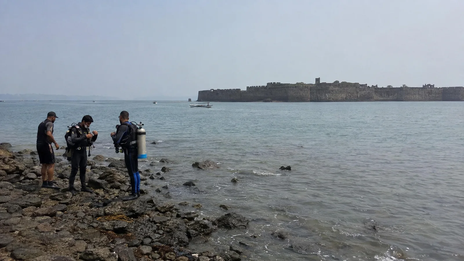 Divers standing on a rocky shore with a coastal fort offshore.