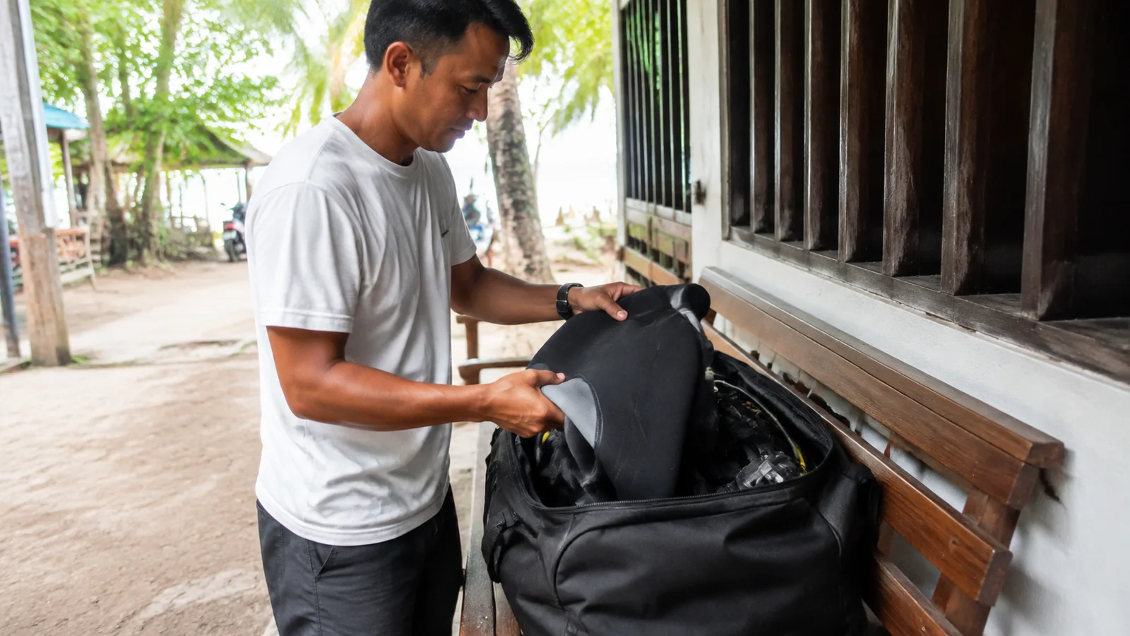 Man packing diving equipment into a bag beside a wooden building.