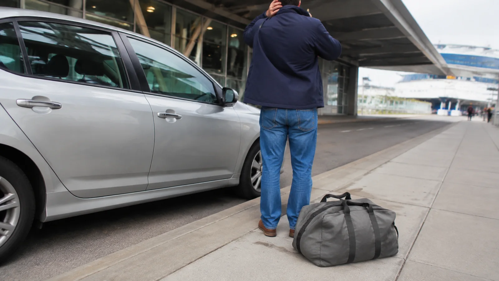 Traveler waiting beside a car with a duffel bag at a terminal.