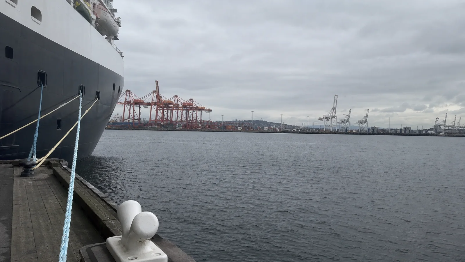 Cruise ship's mooring lines beside a harbor with container cranes.