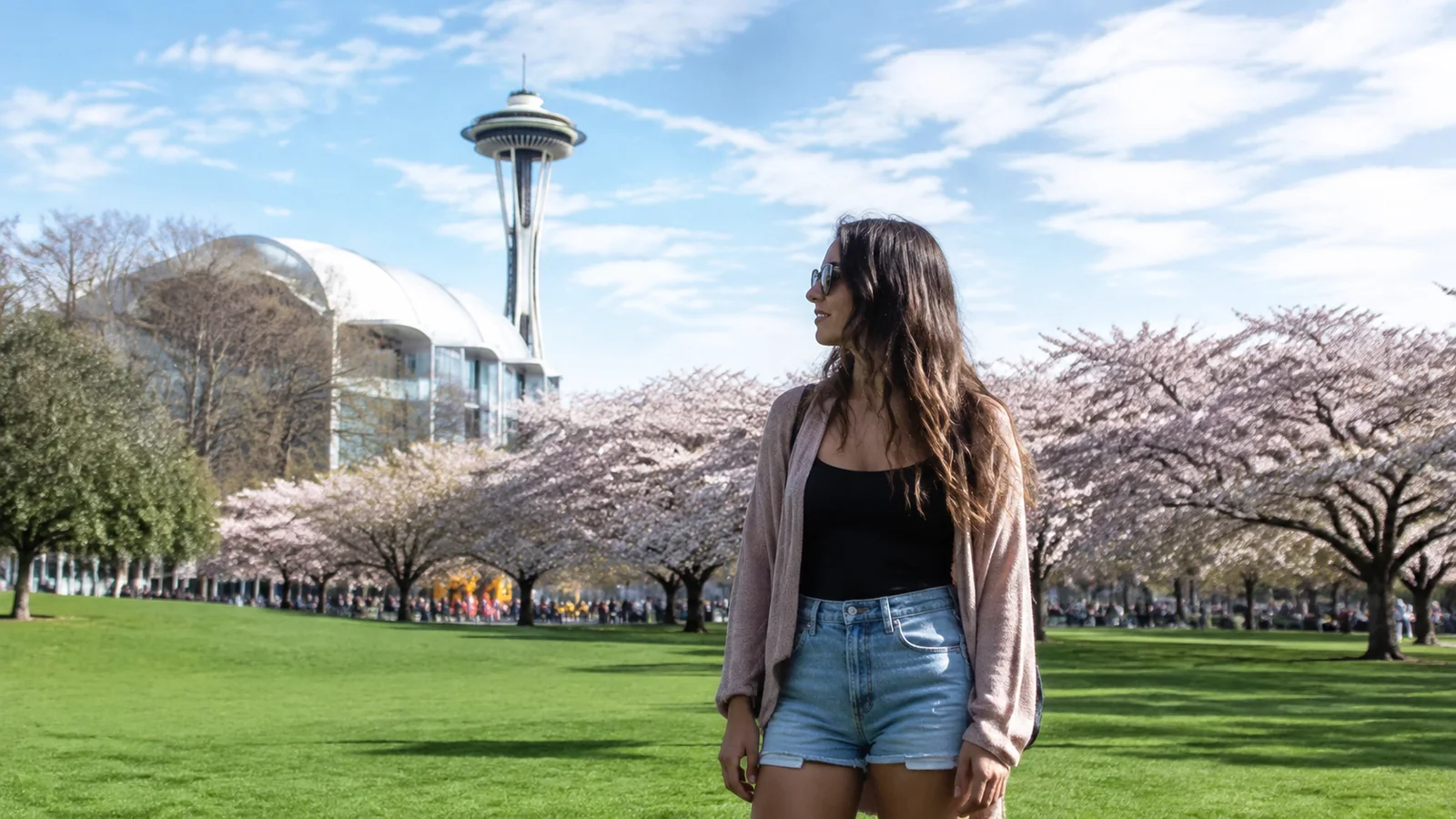 Woman standing in a park with flowering trees and a tall observation tower.