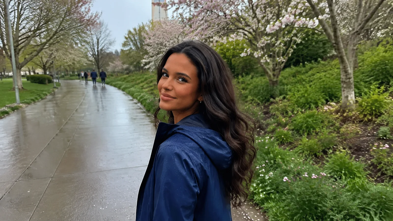 Woman in a blue rain jacket on a wet path beneath flowering trees.