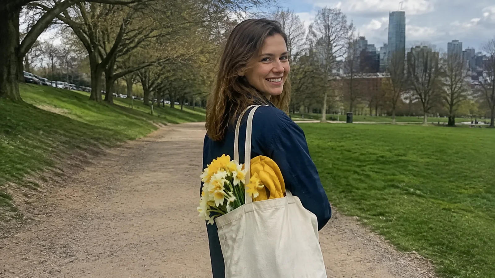 Woman carrying a tote filled with yellow flowers along a park path.