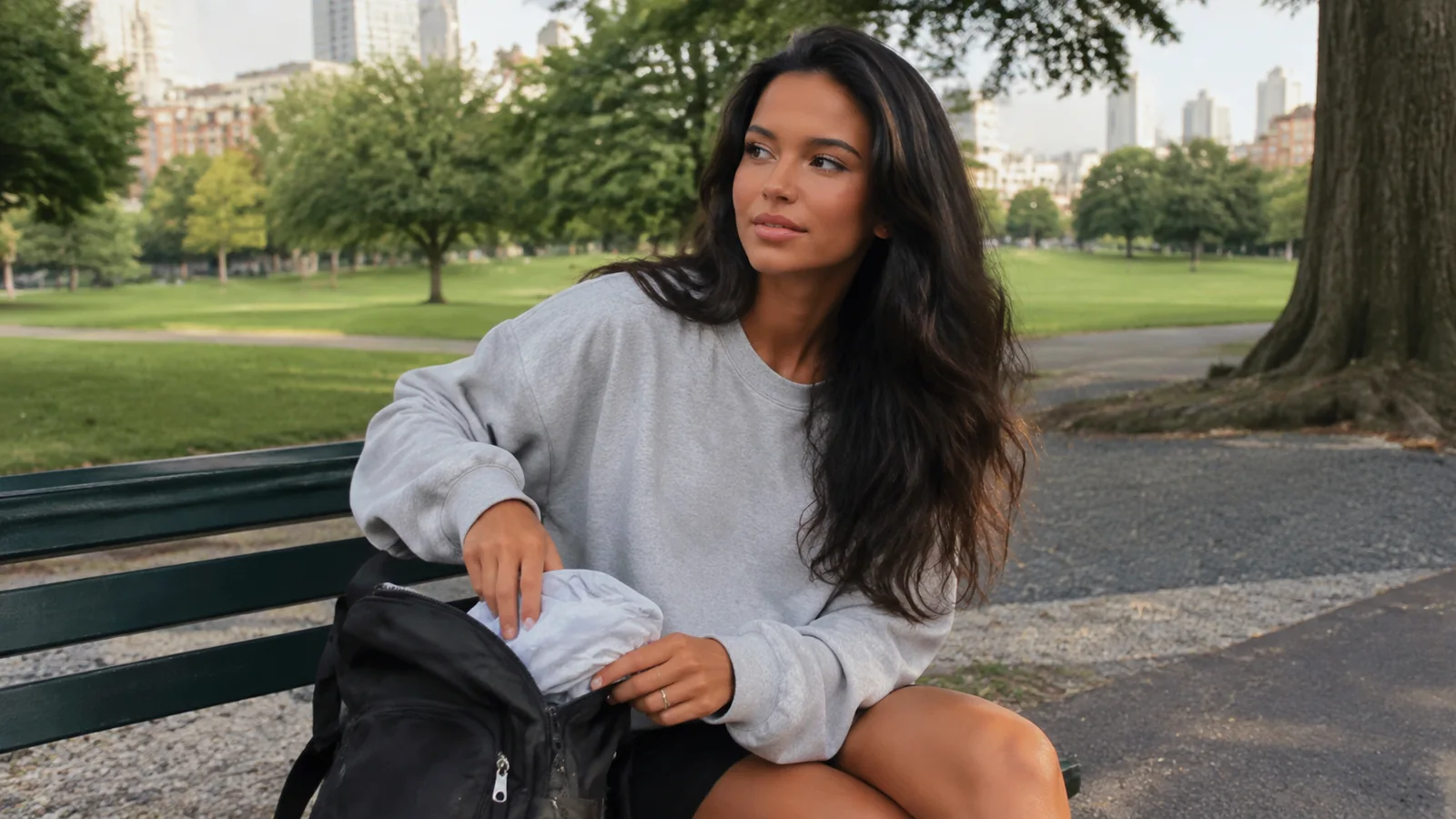 Woman opening a black bag while seated on a green park bench.