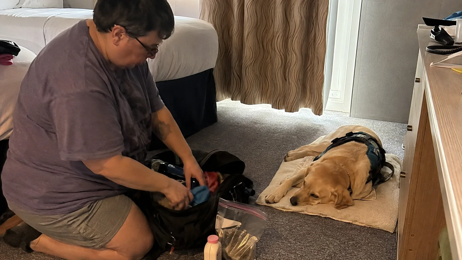 Traveler packing a bag while a harnessed dog rests nearby.