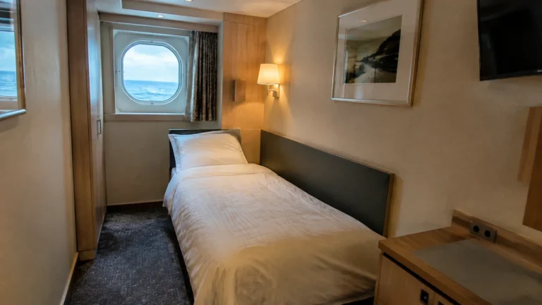 Narrow ship cabin with a single bed beneath a round ocean-facing window.
