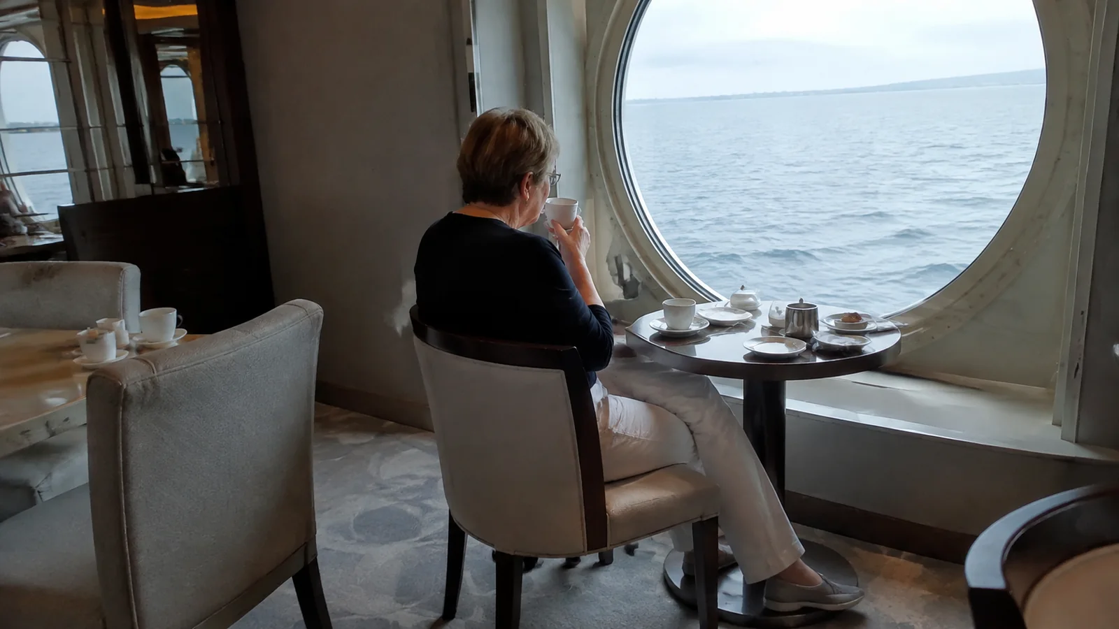 Woman sitting with a cup beside a large round window overlooking the sea.