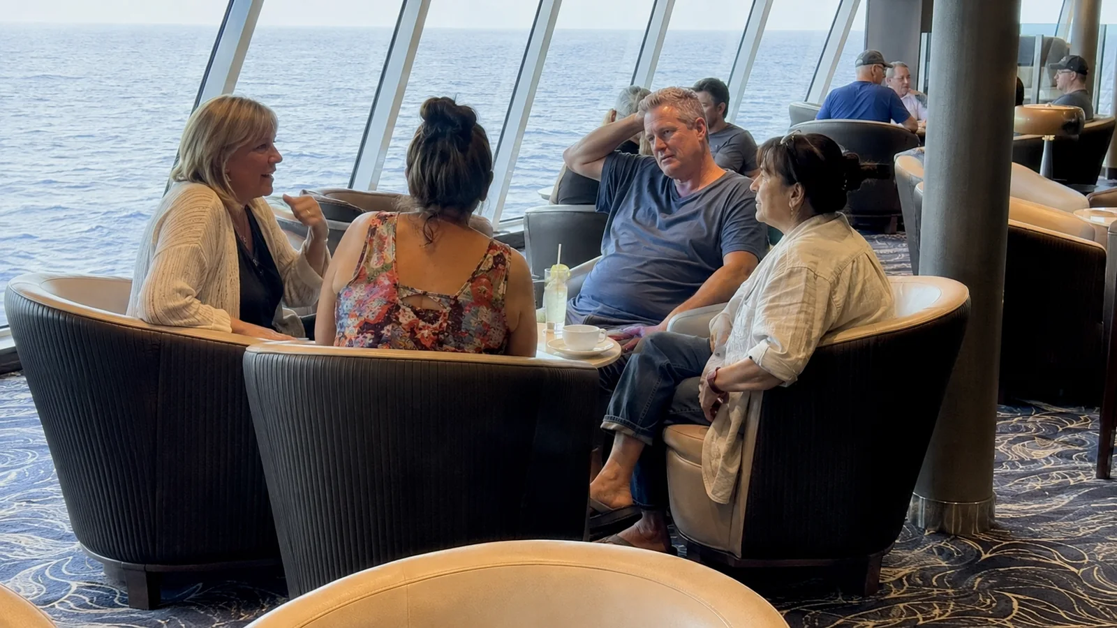 Four passengers chatting around a small table in an ocean-view lounge.