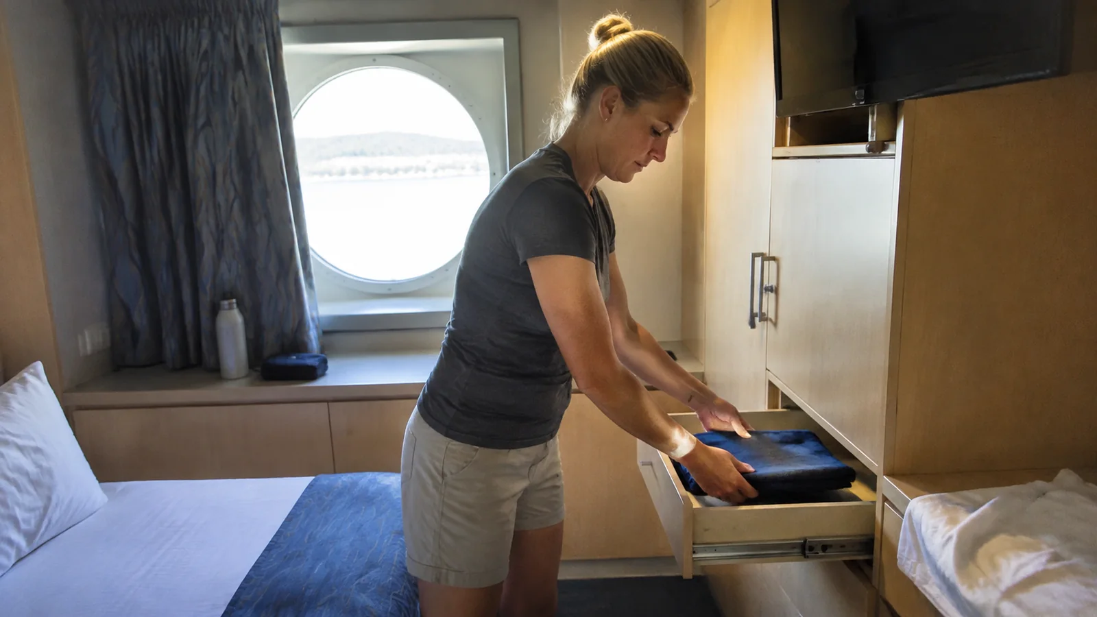 Woman placing folded clothing into a drawer in a compact ship cabin.