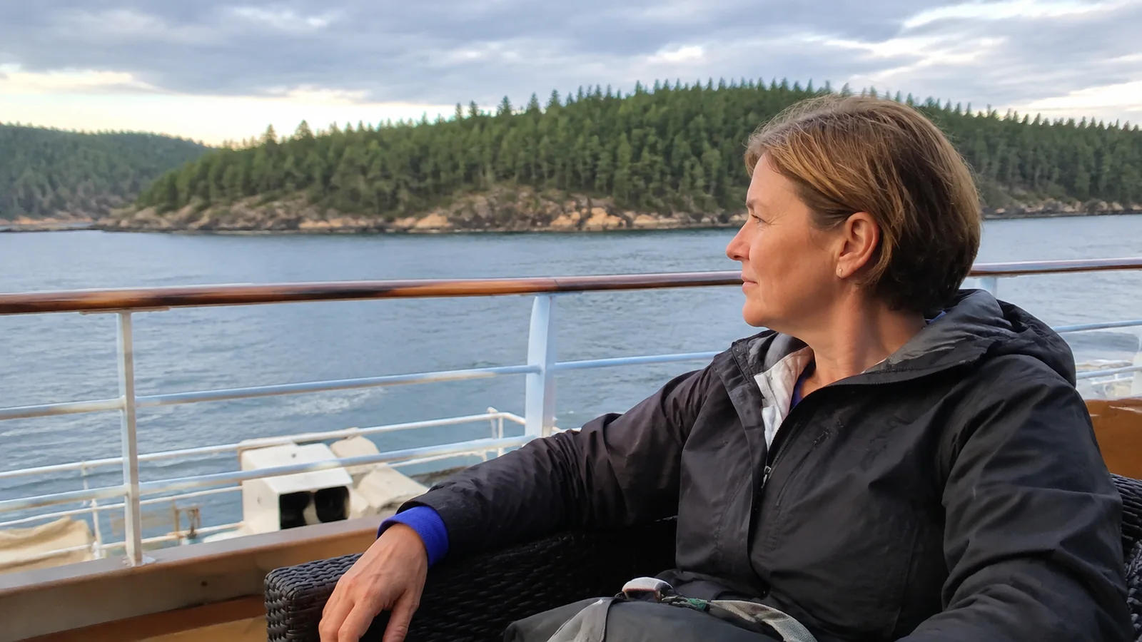 Passenger rests beside a boat railing overlooking a forested shoreline.