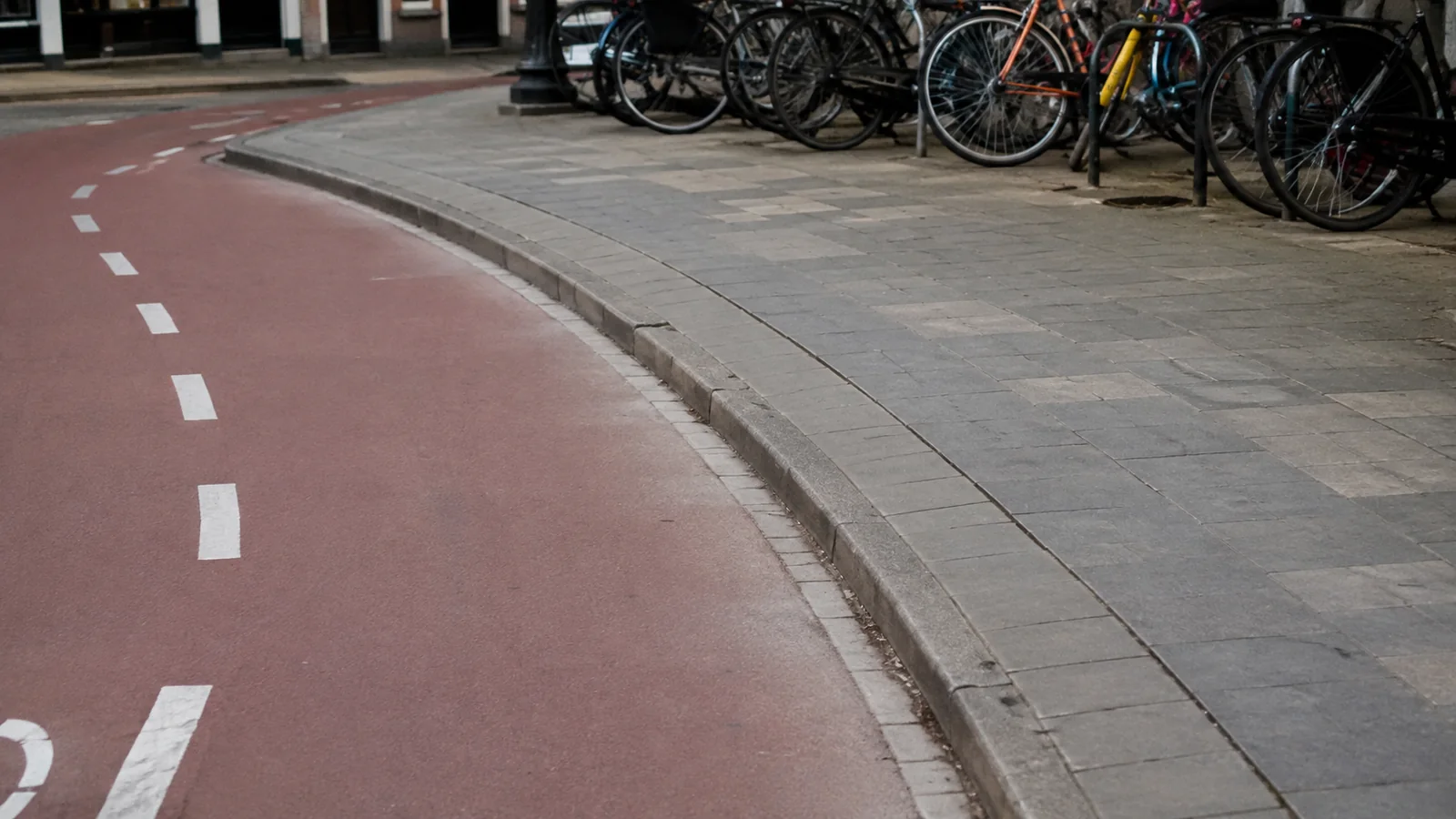 Red bicycle lane curves beside a sidewalk lined with parked bicycles.