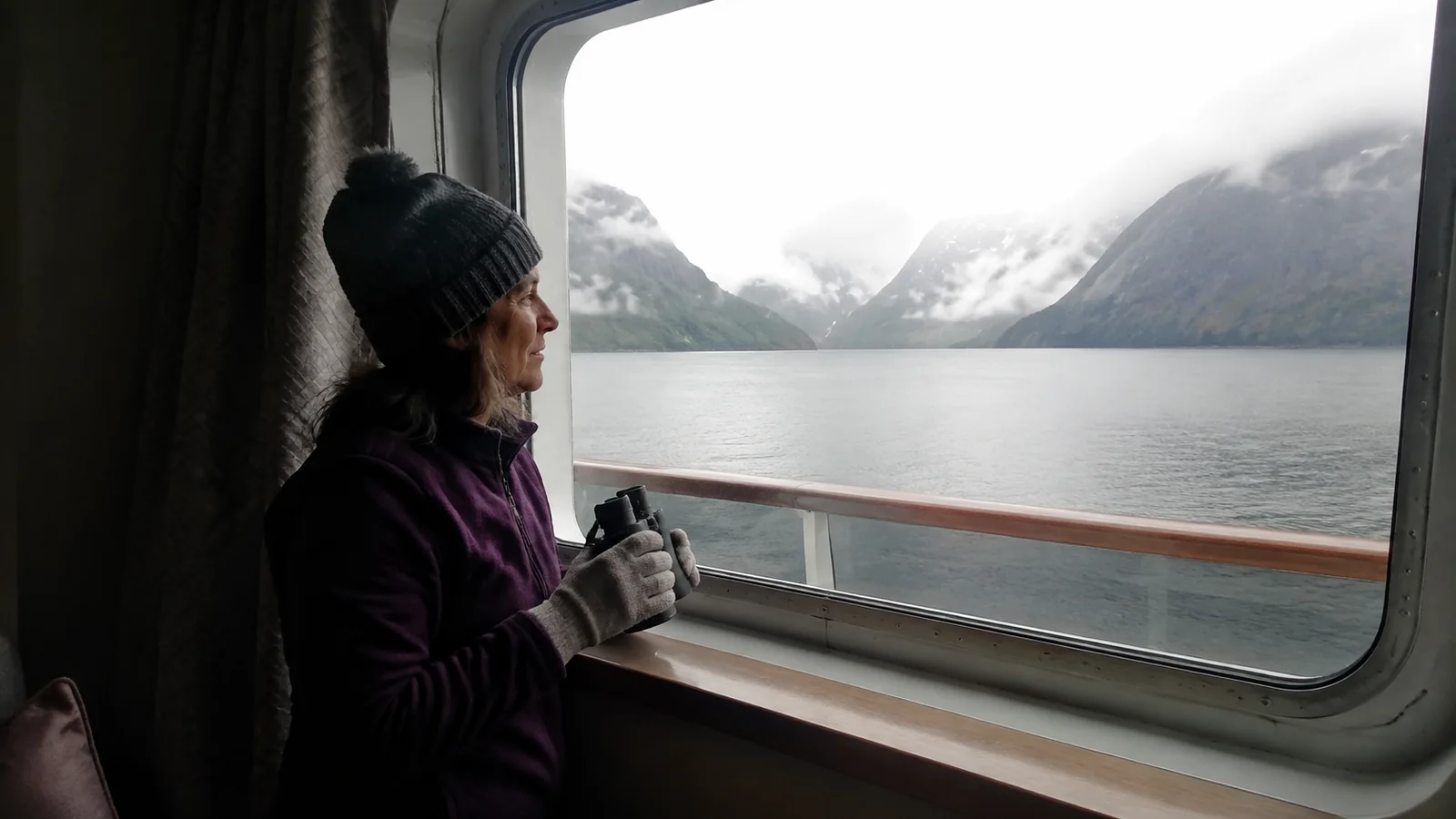 Traveler holding binoculars beside a ship window overlooking steep mountains.