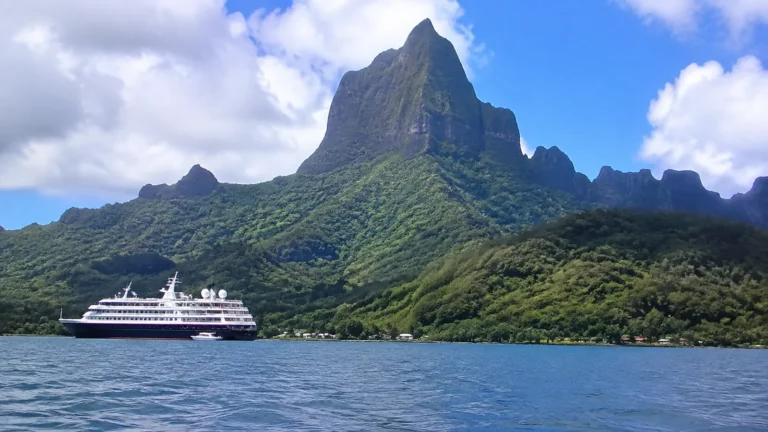Small cruise ship beneath a towering green tropical mountain.