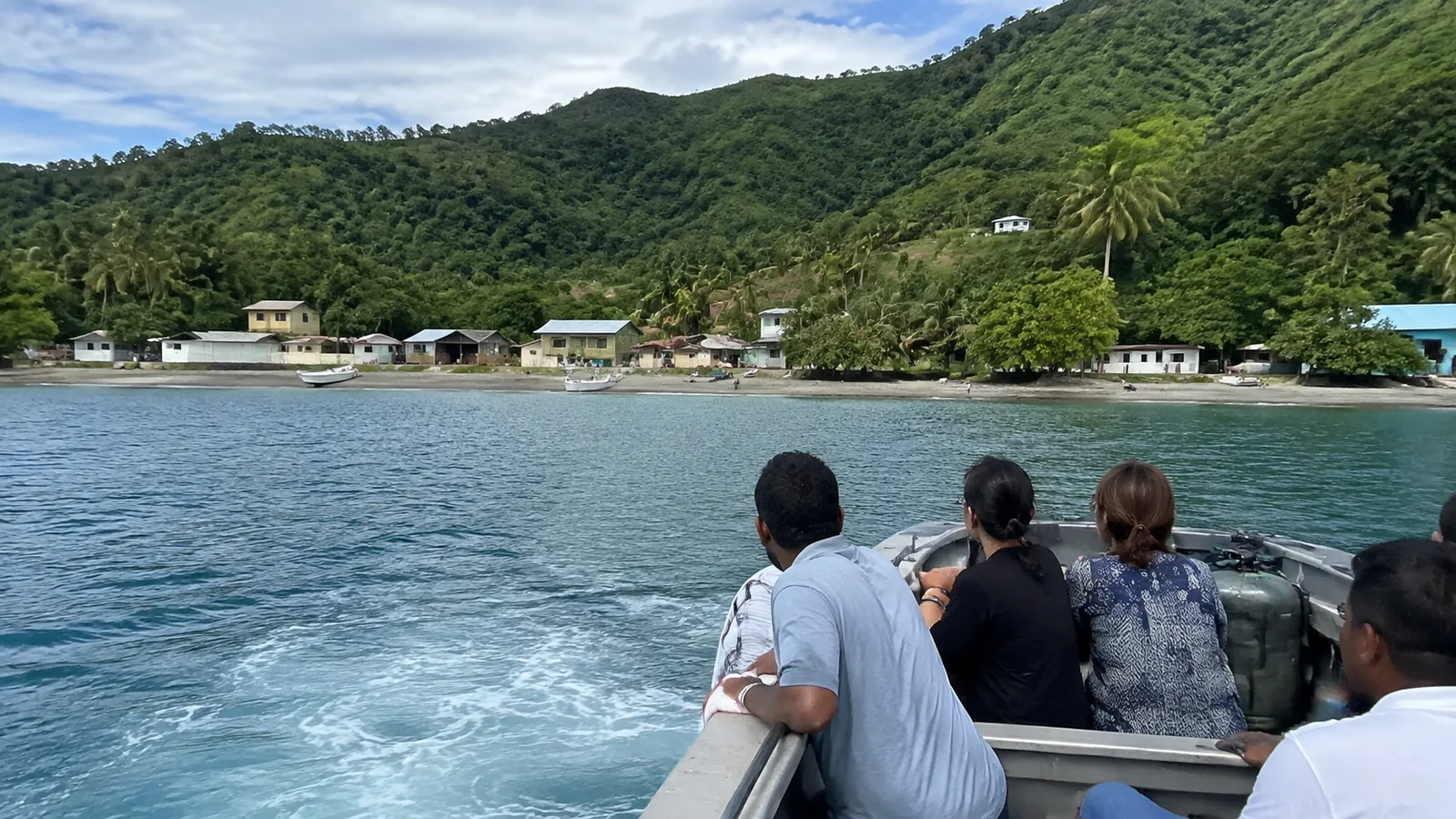 Passengers on a small boat approaching a village below green hills.