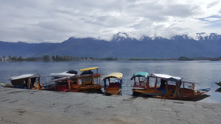 Colorful covered boats moored beside a lake beneath cloud-covered mountains.