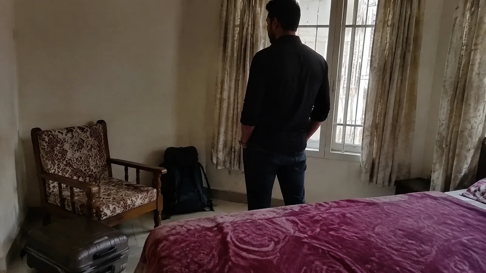Traveler standing beside a window and packed bag in a modest bedroom.