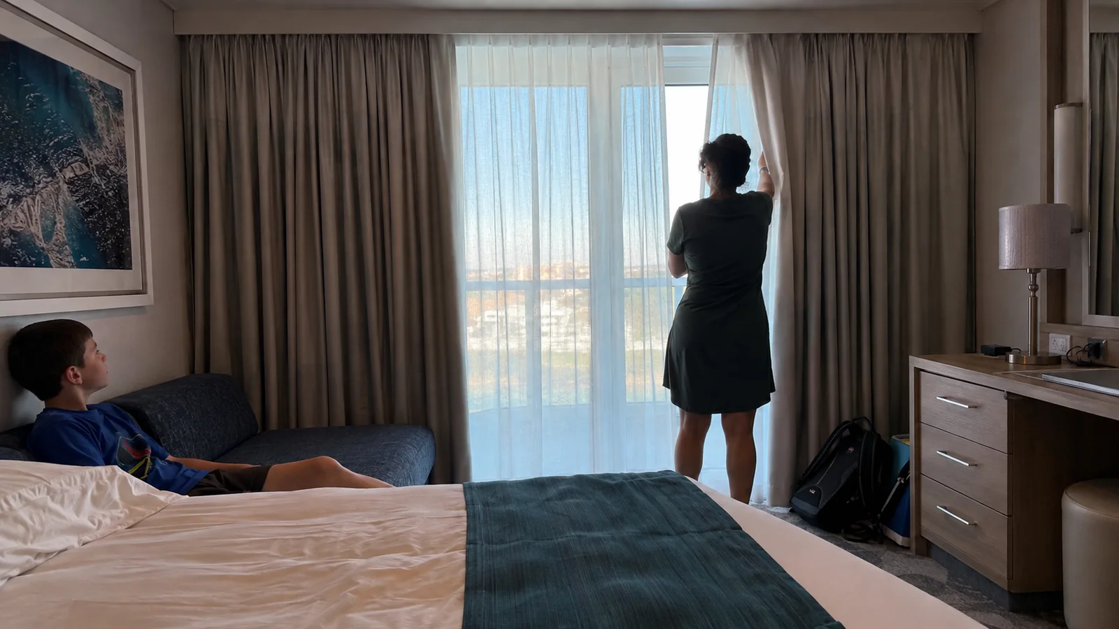 Woman adjusts stateroom curtains while a man rests on the sofa.