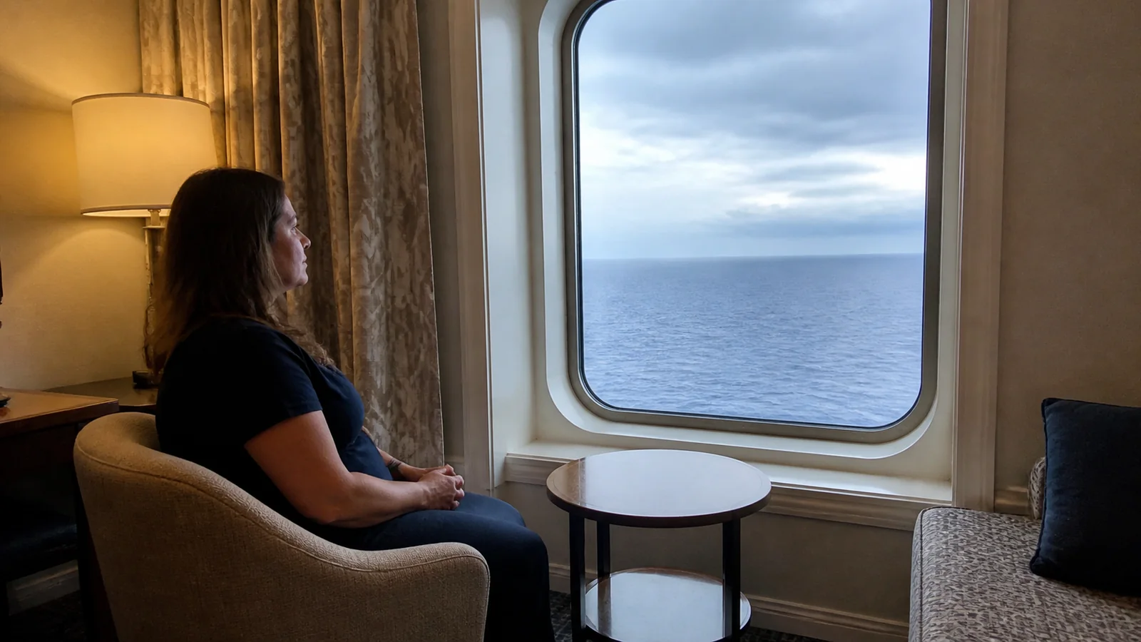 Woman sits beside a large stateroom window looking out at the ocean.