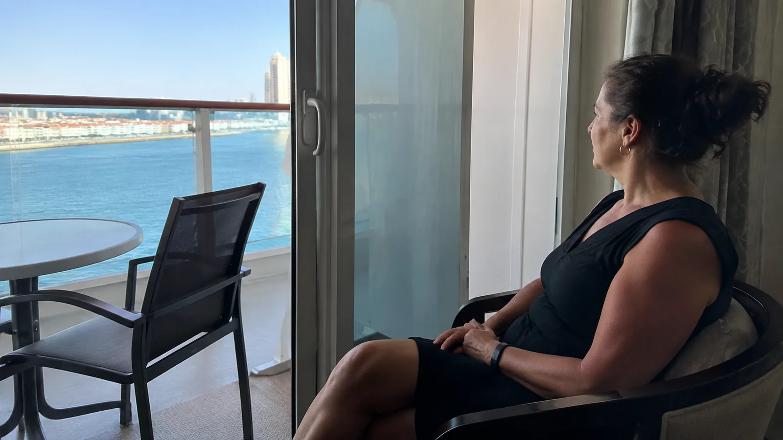 Woman sits inside a stateroom beside an open balcony doorway.