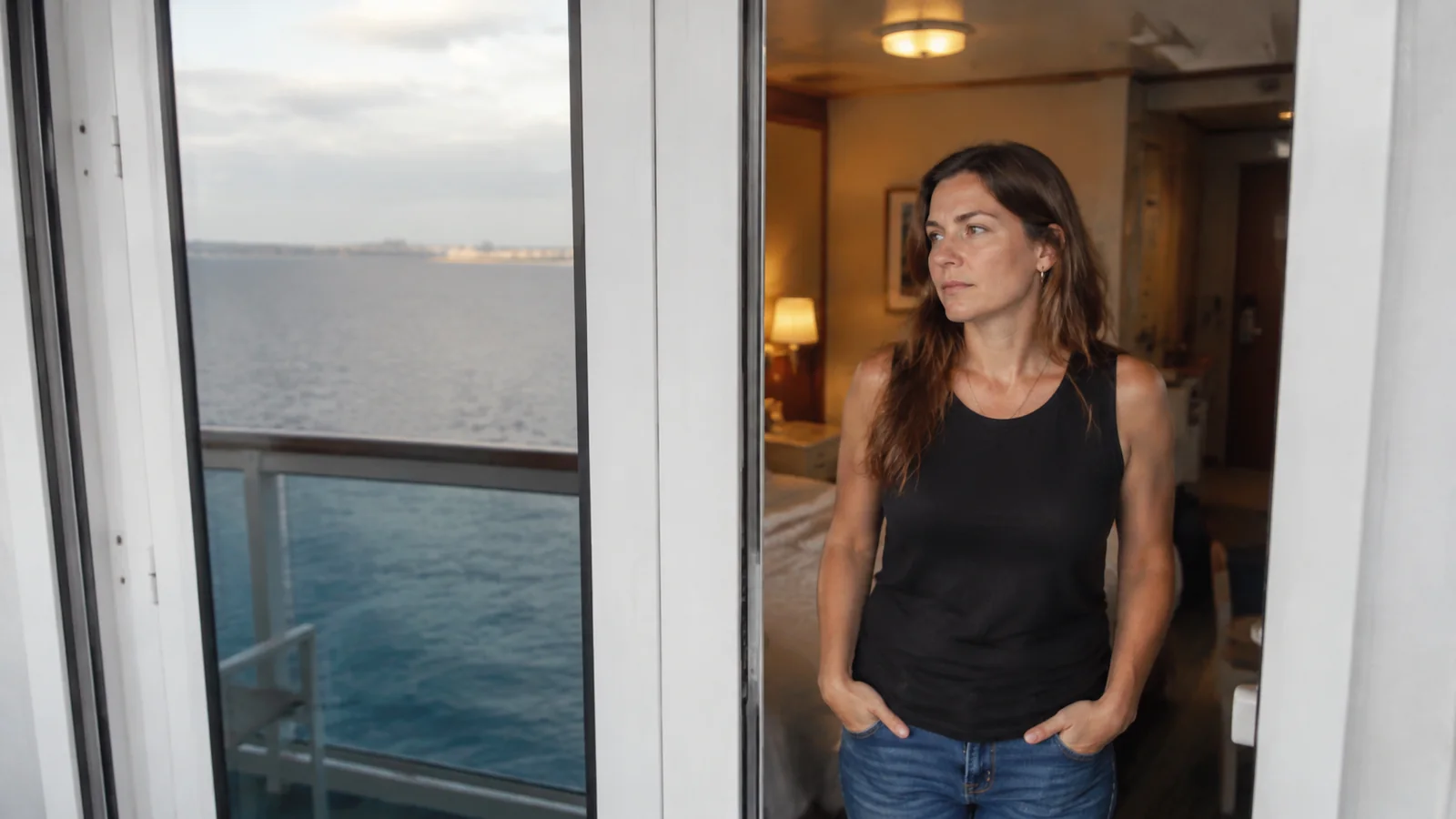 Woman stands beside glass doors looking toward the water.